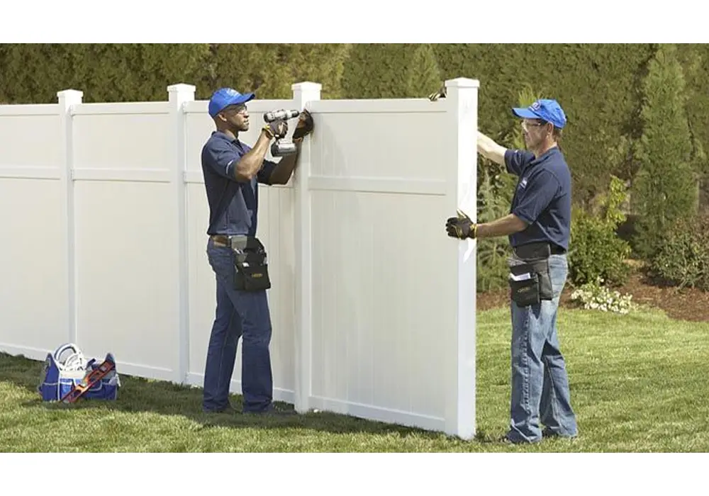 Two contractors performing new fence installation using power tools to secure white vinyl panels in backyard.
