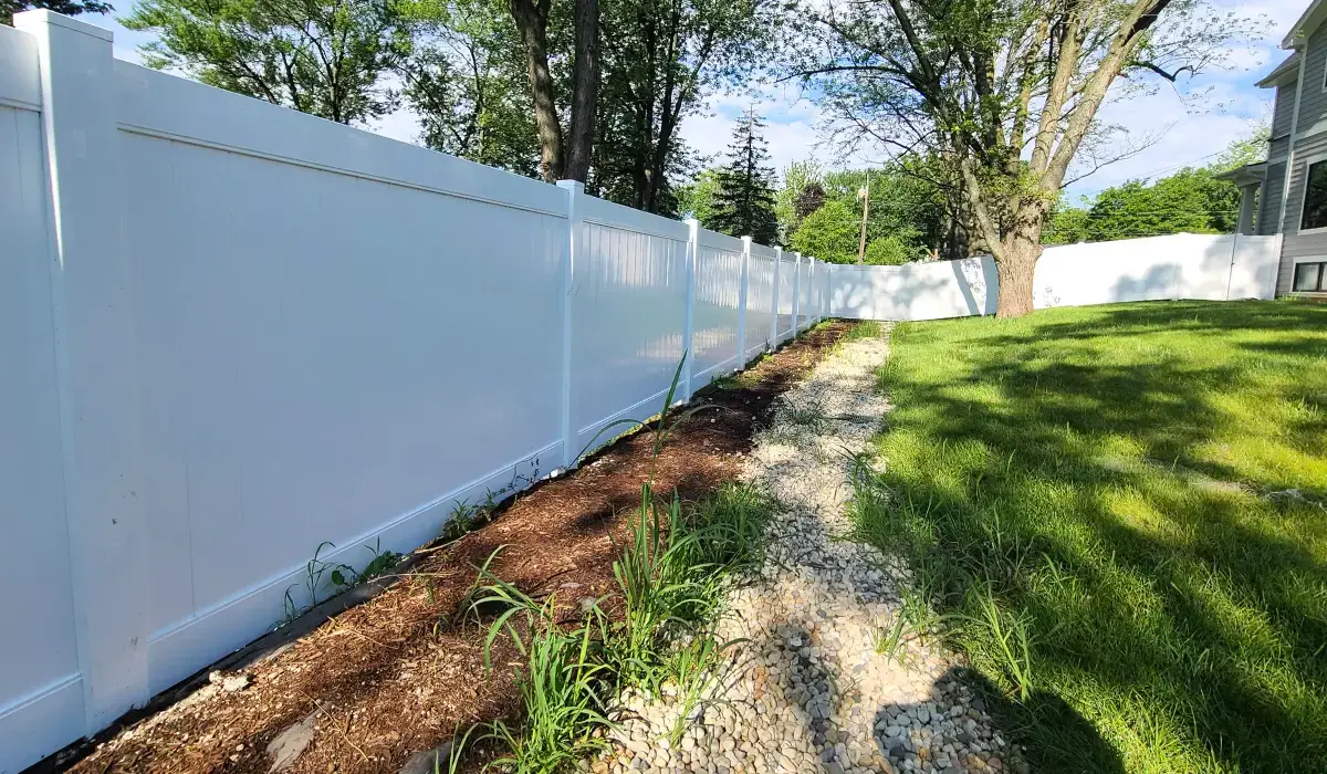 A white vinyl privacy fence runs along a backyard with a gravel pathway, green grass, and tall trees.
