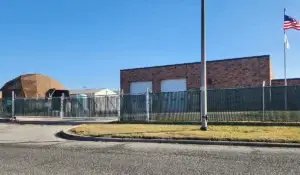 A chain-link commercial fence with privacy slats encloses an industrial facility with brick buildings and a security gate.