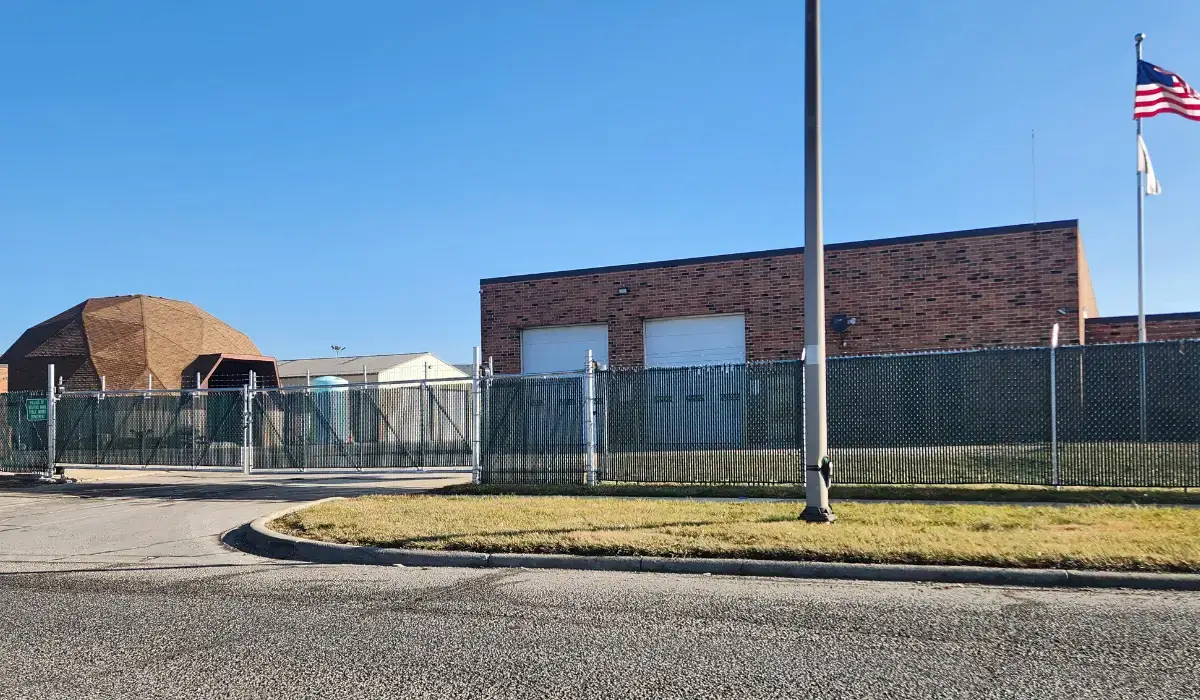 A chain-link commercial fence with privacy slats encloses an industrial facility with brick buildings and a security gate.
